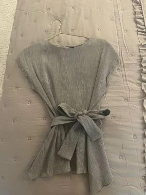 Grå omlottopp med knytband - Säljer en snygg grå omlottopp med korta ärmar och breda knytband i midjan. Toppen har en lätt ribbad struktur och rund halsringning. Perfekt att styla med jeans eller kjol för en trendig look.