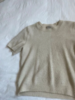 Beige stickad t-shirt från H&M - Kortärmad stickad t-shirt i mjuk beige ullmix från H&M. Tröjan har rund halsringning och ribbade kanter vid ärmslut och nederkant. Perfekt för lager på lager och en clean, minimalistisk look.