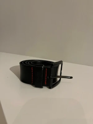 Svart skärp från Hugo med röd söm - Snyggt svart bälte från Hugo i skinn med coola röda sömmar längs kanterna. Klassisk metallspänne i svart finish och diskret Hugo-logga på hällan. Perfekt accessoar för att lyfta din outfit.