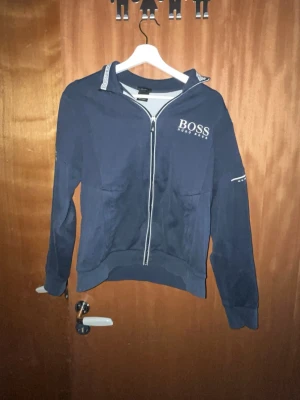 Marinblå zip-hoodie från Hugo Boss - Snygg marinblå hoodie från Hugo Boss med dragkedja framtill och vita detaljer längs ärmarna. Klassisk logga på bröstet och ribbade muddar vid ärmslut och midja. Perfekt för en stilren och avslappnad look.