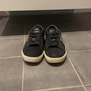 Svarta Saint Laurent sneakers - Svarta sneakers från Saint Laurent med vit logga på sidan. Skorna har en klassisk låg siluett, svarta skosnören och vit sula. Ovandelen är i mocka och läder vilket ger en lyxig känsla. Perfekta för dig som gillar stilrena och exklusiva sneakers. Slitningarna ska finnas de är inte trasiga.