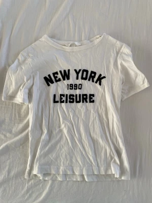 Vit t-shirt med New York-tryck - Vit t-shirt med svart text 'NEW YORK 1990 LEISURE' på bröstet. Klassisk rund halsringning och korta ärmar. Perfekt för en avslappnad och trendig stil. Från hm 😁