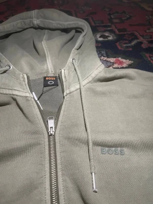 Grå zip hoodie från BOSS - Snygg grå hoodie från BOSS med dragkedja framtill och justerbar huva med snören. Diskret BOSS-logga broderad på bröstet. Tillverkad i mjukt bomullsmaterial som känns skönt mot huden. Perfekt för en avslappnad och stilren look.