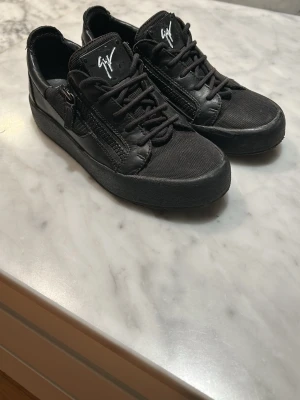 Skor Giuseppe Zanotti - Snygga helsvarta sneakers från Giuseppe Zanotti med dragkedjedetaljer på sidorna och vit logga på plösen. Skorna har en mix av läder och textilmaterial samt klassisk låg siluett med snörning. Perfekta för dig som gillar stilrena och trendiga skor.