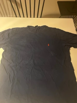 Mörkblå t-shirt från Ralph Lauren - En klassisk mörkblå t-shirt från Ralph Lauren med den ikoniska röda polospelaren broderad på bröstet. T-shirten har rund hals och är tillverkad i mjuk bomull. Enkel och stilren design som funkar till det mesta.