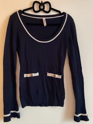 Marinblå långärmad topp med volang på ärmar - Säljer en marinblå långärmad topp från H&M med vita ribbade detaljer vid halsringning, ärmslut och dekorativa fickor framtill. Toppen har volangkant på ärmarna och guldknappar på fickorna för en extra touch. 