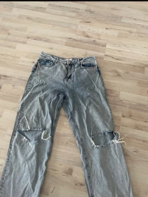Grå slitna jeans från NA-KD - Säljer ett par grå jeans från NA-KD med raka ben och stora slitningar över båda knäna. Jeansen har klassisk femficksmodell och är tillverkade i denim. Perfekta för en avslappnad och trendig look.