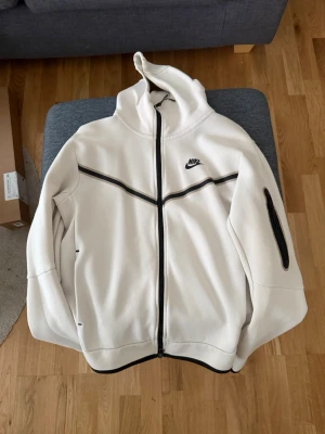 Vit Nike Tech - Snygg vit hoodie från Nike med svarta dragkedjor och detaljer. Har huva, hel dragkedja framtill och en extra ficka med dragkedja på ena ärmen. Perfekt för en sportig och avslappnad stil. Inga märken eller något trasigt, som ny.