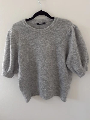 Grå stickad tröja med puffärm - Mysig grå stickad tröja från Gina Tricot med korta puffärmar och rund halsringning. Tröjan har en mjuk och fluffig känsla, ribbstickade muddar och avslappnad passform. Perfekt för lager-på-lager och chill dagar.