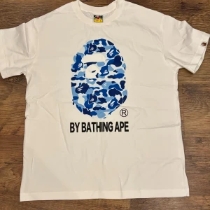 Vit BAPE T-shirt BLÅ logga  - Snygg vit t-shirt från BAPE med det klassiska apansikte-trycket i blått camomönster på bröstet och texten 'BY BATHING APE' under. Perfekt för dig som gillar streetwear och vill sticka ut med en ikonisk design.