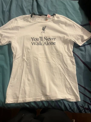 Vit LFC t-shirt med tryck - Vit t-shirt från Liverpool FC med klubbens logga och texten 'You'll Never Walk Alone' tryckt på bröstet. Klassisk passform med korta ärmar och rund hals. Perfekt för dig som vill visa ditt stöd för laget.