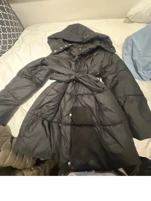 Vero Moda medel lång dam jacka  - Snygg svart dunjacka med huva och ett brett knytband i midjan som ger en markerad siluett. Jackan har långa ärmar, tryckknappar framtill och quiltad design. Perfekt för kalla vinterdagar och riktigt varm.