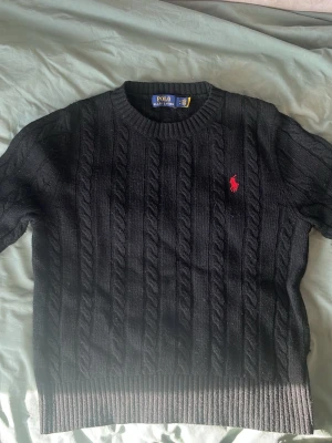 Ralph lauren stickad svart  - Kabelstickad svart tröja från Polo Ralph Lauren med rund halsringning och röd broderad logga på bröstet. Tröjan har ribbade muddar vid ärmslut och nederkant. Klassisk och tidlös design i mjukt material.