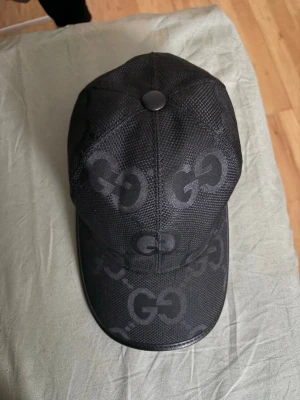 Svart keps från Gucci med GG-mönster - Säljer en svart keps från Gucci med det klassiska GG-mönstret i ton-i-ton. Kepsen har böjd skärm och är tillverkad i ett mesh-liknande material med detaljer i konstläder runt skärmen. Perfekt för dig som vill ha en stilren och lyxig accessoar.