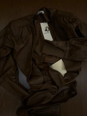 Brun topp från Adoore - Snygg brun omlottopp i läder från Adoore i klassisk modell med axelpartier och knappar vid ärmslut. 