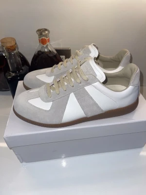 Maison Margiela - Snygga sneakers i vitt läder och ljusgrå mocka med beige snören och brun gummisula. Klassisk och stilren design som passar till det mesta. Skorna har lågt skaft och en tidlös look inspirerad av retro-modeller.