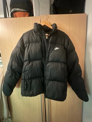 Svart pufferjacka från Nike - Säljer en svart pufferjacka från Nike med hög krage och dragkedja framtill. Jackan har två sidofickor med dragkedja och den klassiska Nike-loggan broderad i vitt på bröstet. Perfekt för kalla dagar och riktigt skön passform. Storlek xs funkar för dig som har S