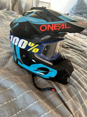 Svart och blå crosshjälm O'Neal - Säljer en svart och blå crosshjälm från O'Neal med röd logga och cool design. Hjälmen har ventilationshål, justerbart visir och mjuk, vadderad insida i rött och grått. Medföljer blåa 100% goggles med bred elastisk rem.