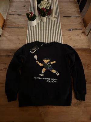 Mörkblå sweatshirt Polo Bear Ralph Lauren - Mörkblå sweatshirt från Ralph Lauren med ikoniska Polo Bear-trycket där björnen spelar tennis. Rund halsringning och ribbade muddar. Perfekt för dig som gillar sportig och trendig stil.