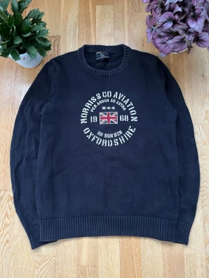Svart stickad tröja Morris Oxfordshire - Svart stickad tröja från Morris med rund halsringning och tryck framtill med brittisk flagga och texten 'Morris & Co Aviation Oxfordshire'. Tröjan har ribbade muddar vid ärmslut och nederkant. Perfekt för en stilren och avslappnad look.