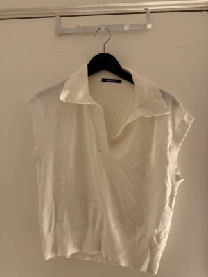 Vit ärmlös topp med krage - Stilren vit topp från Gina Tricot med klassisk krage och djup v-ringning. Modellen är ärmlös och har en mjuk, stickad känsla som passar perfekt till jeans eller kjol.
