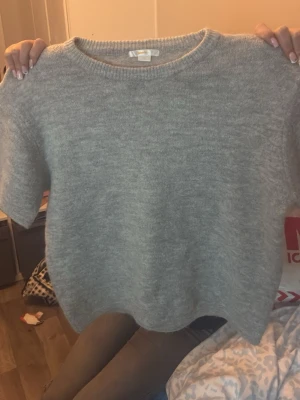 Grå stickad tröja med kort ärm - Mysig grå stickad tröja med korta ärmar och rund halsringning. Tröjan har en enkel och stilren design, perfekt att matcha med jeans eller kjol. Materialet ser ut att vara mjukt och fluffigt, vilket ger en avslappnad vibe.