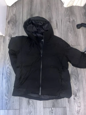 Uniqlo jacka  - Säljer en svart pufferjacka med huva och dragkedja framtill. Jackan har en klassisk boxig passform och är vadderad för extra värme. Perfekt för kalla vinterdagar och riktigt snygg till streetwear-stilen.