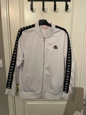 Vit Kappa track tröja med svarta detaljer - Snygg vit track tröja från Kappa med svarta ränder och loggor längs ärmarna. Tröjan har dragkedja framtill, ribbade muddar och klassisk Kappa-logga på bröstet. Perfekt för en sportig och trendig look.