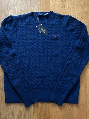 Mörkblå kabelstickad tröja Polo Ralph Lauren - storlek S - Mörkblå kabelstickad tröja från Polo Ralph Lauren med rund halsringning och röd broderad logga på bröstet. Tröjan har långa ärmar och ribbade muddar vid ärmslut och nederkant. Perfekt för dig som gillar klassisk och stilren look. Storlek - S