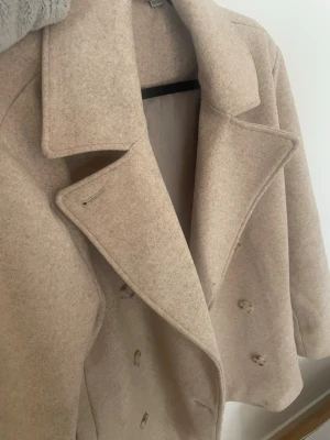 Beige ullkappa från Zink - Stilren beige kappa från Zink i storlek L (44/46). Kappan har dubbelknäppning, bred krage och är tillverkad i ett mjukt ullmaterial. Perfekt för dig som vill ha en klassisk och tidlös look med en modern twist.