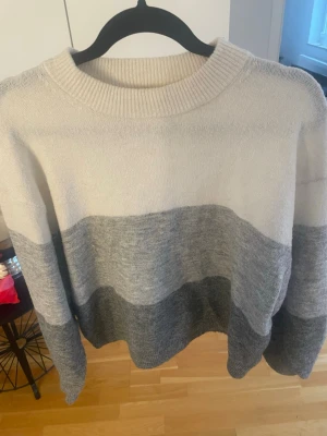 Gråvit stickad tröja från H&M - Säljer en stickad tröja från H&M i storlek M. Tröjan har breda blockränder i vitt och olika nyanser av grått, rund halsringning och långa ärmar. Perfekt för dig som gillar en enkel men snygg stil.