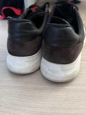 Svarta och vita sneakers från Axel Arigato - Snygga sneakers från Axel Arigato i svart mocka och läder med vit sula och vit sidopanel. Klassisk låg modell med svarta skosnören och diskret Arigato-logga på sidan och hälen. Perfekta för en clean och stilren look.