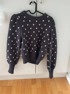 Mörkgrå stickad tröja med vita prickar - Mysig mörkgrå stickad tröja med vita prickar och puffiga ärmar. Tröjan har rund halsringning och ribbade muddar vid ärmslut och nederkant. Perfekt för dig som gillar en söt och trendig look.