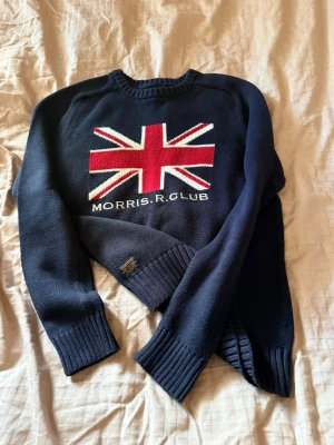Mörkblå stickad tröja Morris R Club - Snygg mörkblå stickad tröja från Morris R Club med stor Union Jack-flagga i rött, vitt och blått på bröstet. Tröjan har rund halsringning och ribbade muddar vid ärmslut och nederkant. Perfekt för dig som gillar klassisk brittisk stil.