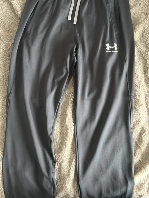 Gråa mjukisbyxor från Under Armour - Snygga grå mjukisbyxor från Under Armour med vit logga på låret och vita dragsnören i midjan. Tillverkade i ett mjukt och stretchigt material som känns skönt mot huden. Perfekta för chill eller träning.