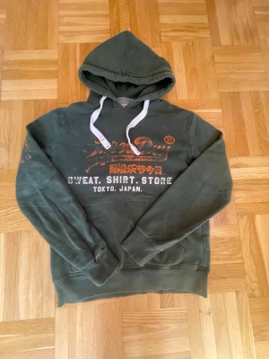 Grön Superdry hoodie - Mörkgrön hoodie från Superdry med stort vintage-tryck i orange och vitt på bröstet. Klassisk huva med vita snören och känguruficka framtill. Skön och avslappnad passform, perfekt till jeans eller joggers. Textdetaljer på ärmen och bröstet för extra stil. Passar ca 175 cm.