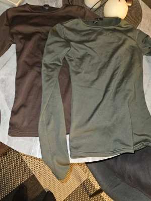 Två långärmade tröjor från Amisu - Säljer två stilrena långärmade tröjor från Amisu i storlek S. En är olivgrön och den andra är mörkbrun. Båda har rund halsringning och är tillverkade i ett mjukt, stretchigt material som sitter snyggt på kroppen. Perfekta att matcha med jeans eller kjol.