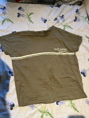 Olivgrön t-shirt med vit rand - Snygg olivgrön t-shirt med vit horisontell rand och texten 'SAN LEGO' på bröstet. T-shirten har rund hals och korta ärmar, perfekt för en avslappnad stil. Tillverkad i mjuk bomull för skön komfort.