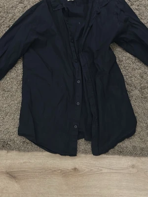 Mörkblå skjorta med knappar - En stilren mörkblå skjorta med långa ärmar och klassisk krage. Skjortan har knäppning framtill och är tillverkad i ett mjukt tyg som känns bekvämt mot huden. Perfekt för dig som gillar en enkel men snygg look. Från lager 157, bara använd ett fåtal gånger.