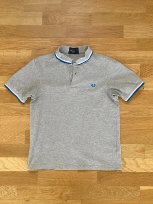 Grå pikétröja från Fred Perry - Klassisk grå pikétröja från Fred Perry med blå och vit rand på krage och ärmslut. Tröjan har korta ärmar, knappslå framtill och den ikoniska Fred Perry-loggan broderad på bröstet. Tillverkad i mjuk bomull för en bekväm känsla. Size m but fits more like s