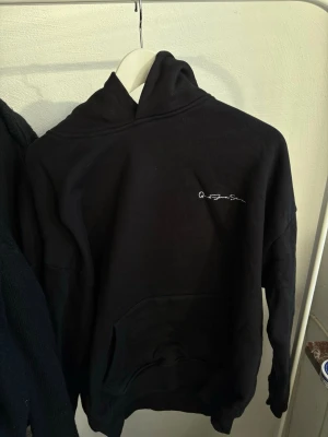 Svart hoodie Que Sera Sera  - Svart hoodie med vit text 'Que Sera Sera' tryckt på ryggen och mindre logga på bröstet.