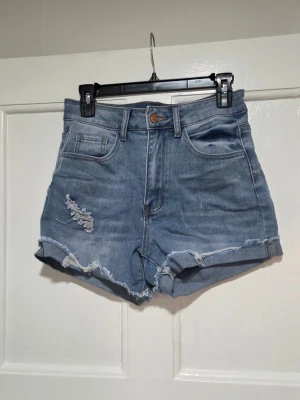 Blå högmidjade jeansshorts - Nya & oanvända! Snygga blå jeansshorts med hög midja och råa kanter. Shortsen har slitna detaljer både fram och bak samt klassiska fem fickor. Perfekta för en avslappnad och trendig look.