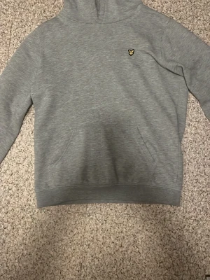 Grå hoodie från Lyle & Scott - Snygg grå hoodie från Lyle & Scott med klassisk gul logga på bröstet. Hoodien har en stor känguruficka framtill och ribbade muddar vid ärmslut och nederkant. Perfekt för en chill och avslappnad stil.