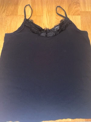 Svart linnetopp med spetskant - Snygg svart linnetopp med tunna axelband och dekorativ spetskant vid urringningen. Materialet känns mjukt och lätt, perfekt för lager på lager eller som den är. Enkel men ändå stilren design som passar till mycket.