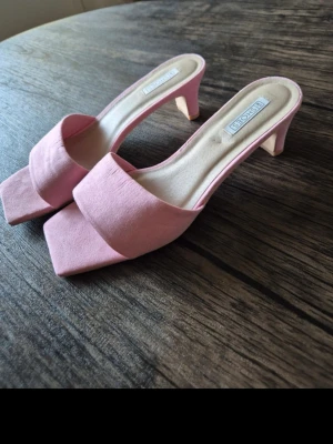 Rosa klackade sandaler från NLY  - Snygga rosa sandaler med klack från Bianco. De har en fyrkantig tå, bred rem över foten och öppen häl. Klacken är medelhög och skon har en mjuk textil på ovandelen. Perfekta för dig som vill ha en trendig och feminin look.
