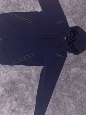 Mörkblå vindjacka från Under Armour  med huva - Stilren mörkblå vindjacka med hel dragkedja framtill och två snedställda fickor med dragkedja. Jackan har en justerbar huva och är tillverkad i ett lätt, vattenavvisande material. Perfekt för blåsiga dagar och enkel att matcha med olika outfits.