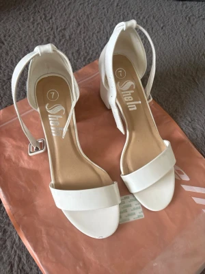 Vita pumps med blockklack från Shein - Säljer ett par stilrena vita pumps från Shein med öppen tå och bred rem över foten. Skorna har en chunky blockklack och justerbart ankelband med silverfärgat spänne. Materialet är konstläder med en slät finish. Perfekta för dig som vill ha en clean och modern look. Lite små märken där framme på ena men inget som syns. 
