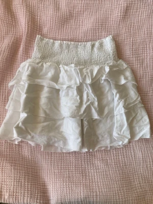 Vit volangkjol med resårmidja - Supersöt vit kortkjol med flera lager volanger och bred smockresår i midjan. Kjolen har en luftig och flowy känsla, perfekt för varma dagar. Materialet känns lätt och skönt mot huden.