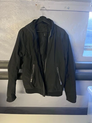 Svart bomberjacka från H&M - Stilren svart bomberjacka från H&M med två fickor framtill som har silvriga dragkedjor. Jackan har klassisk bomberpassform med ribbade muddar vid ärmslut men längre krage. Perfekt för dig som vill ha en enkel men snygg jacka till vardags.