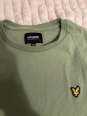 Grön t-shirt från Lyle & Scott - Snygg ljusgrön t-shirt från Lyle & Scott med klassisk rund hals och det ikoniska gula örnlogot på bröstet. Tillverkad i mjuk bomull och har en enkel, stilren design som passar perfekt till jeans eller shorts.